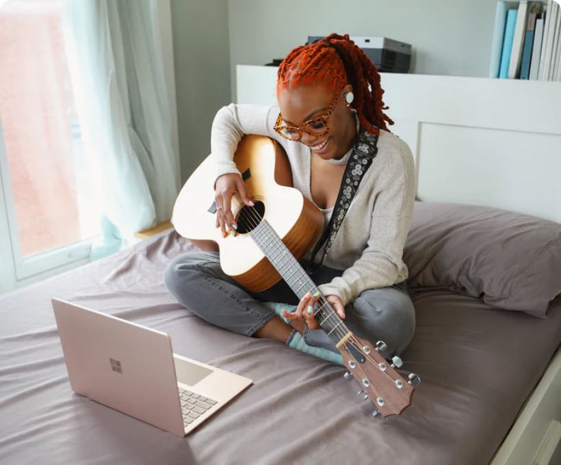 Girl sitting in her bed, in-front of a laptop playing a guitar